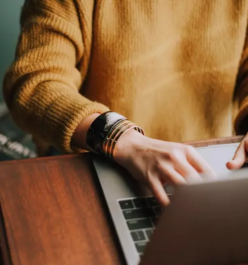 En kvinne med gul strikkegenser og smykker på armen skriver på en bærebar datamaskin.